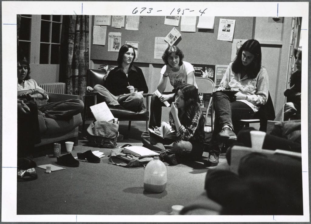 A photo of four women sitting in chairs in a semicircle and one on the floor. Beverages, backpacks, and stacks of papers are scattered on the floor around them. They appear to be writing and talking to each other.